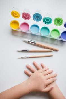 Childs hands with watercolor paints and brushes on a white surface.