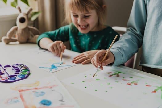 Two cute little sisters or classmates are painting on table with brush. Small girls are laughing. Kids are in kindergarten or at home. Child in living room. Home schooling concept, doing homework