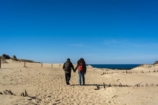 A couple of travelers with a backpack, hand in hand, walks along the sandy Dead Dunes, on the Curonian Spit, on the shore of the Baltic Sea.