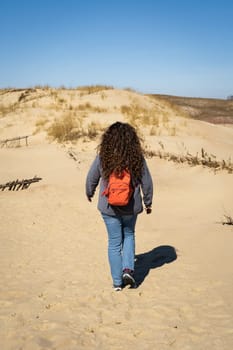 Long-haired curly-haired woman with a backpack walking on sand dunes.