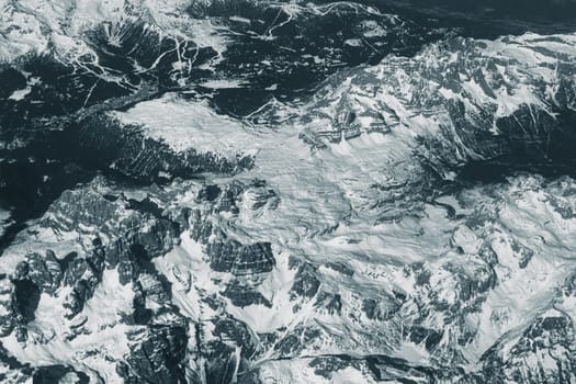 Aerial view of high rocky mountains covered with snow. Alpine landscape. Beauty in nature. Inaccessible mountain peaks. Harsh climate. Looking at the Earth from an airplane window. Aerial view of the land