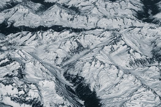 Aerial view of high rocky mountains covered with snow. Alpine landscape. Beauty in nature. Inaccessible mountain peaks. Harsh climate. Looking at the Earth from an airplane window. Aerial view of the land
