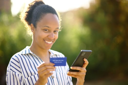 Black woman looking at you holding credit card and phone in a park