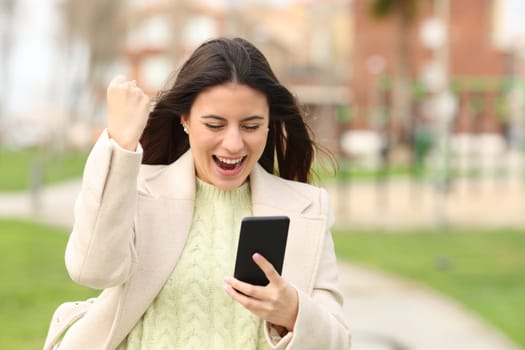 Excited woman checking smart phone content walking in a park in winter