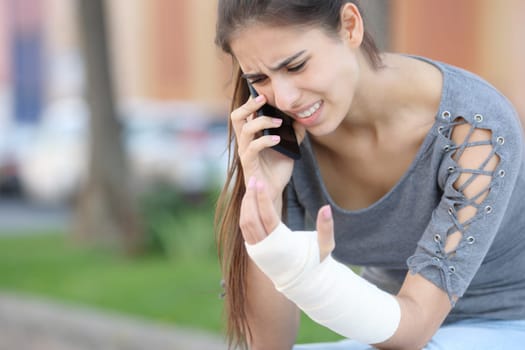 Convalescent woman with bandaged arm calling doctor on phone sitting in the street