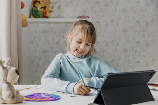 Schooler cute little girl sitting at desk, doing homework using laptop in bedroom, kid studying at home, writing notes, watching tablet. Home education, homeschooling. Child is happy and smiling