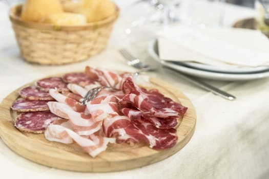 Various appetizer of charcuterie served on a wooden chopping board. On defocused background bread basket, plate, fork.