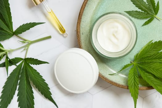 Pipette with oil and opened cream jar with white lid near green cannabis leaves on plate on white marble table top view, mockup. Organic skincare beauty products, natural cosmetics
