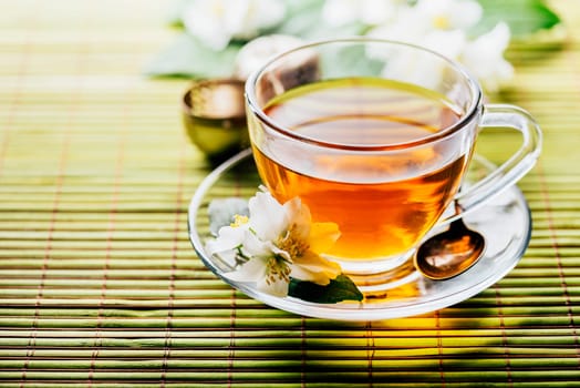 Glass of hot herbal tea on bamboo background closeup. Teacup with organic green tea and fresh jasmine flower on wooden table. Healthy alternative medicine and natural detox beverage.