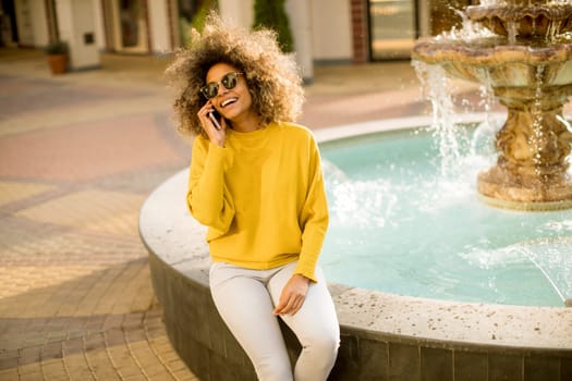 Aftican american young woman sits near fountain and talks on smartphone
