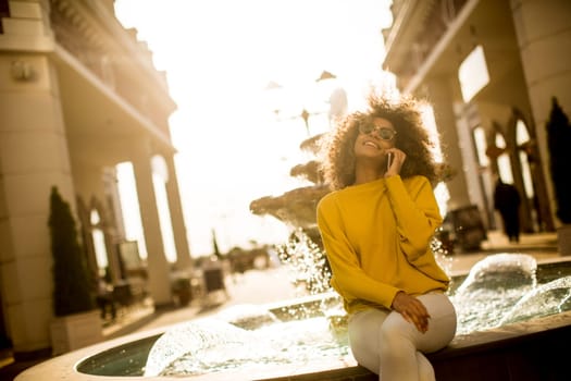 Attractive young woman with curly hair using her touch screen mobile cell phone by the fountain