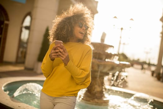 Attractive young woman with curly hair using her touch screen mobile cell phone by the fountain