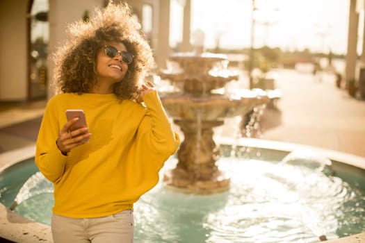 Attractive young woman with curly hair using her touch screen mobile cell phone by the fountain