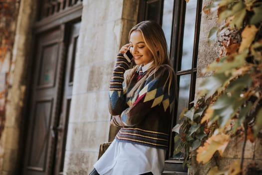 Pretty young woman standing with mobile phone on street at sunny autumn day