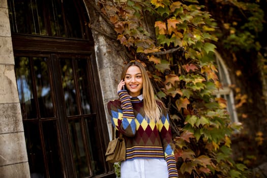 Pretty young woman standing with mobile phone on street at sunny autumn day