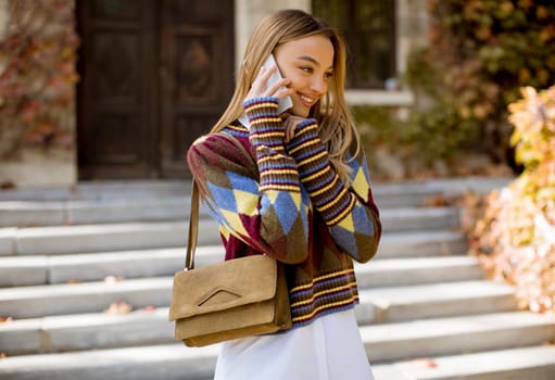 Pretty young woman standing with mobile phone on street at sunny autumn day
