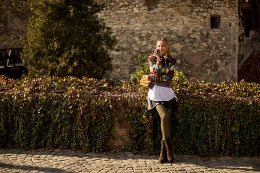 Pretty young woman standing with mobile phone on street at sunny autumn day
