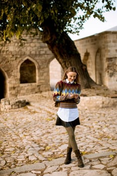 Pretty young woman standing with mobile phone on street at sunny autumn day