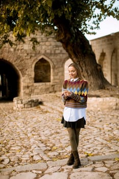 Pretty young woman standing with mobile phone on street at sunny autumn day