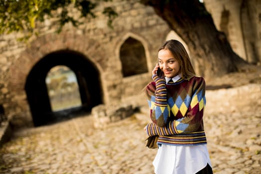 Pretty young woman standing with mobile phone on street at sunny autumn day