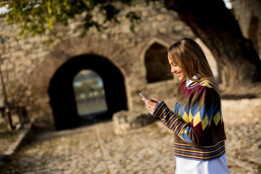 Pretty young woman standing with mobile phone on street at sunny autumn day