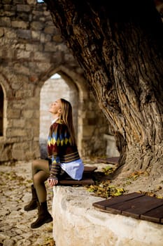 Pretty young woman sitting on the bench watching yellow leaves tree in the autumn park