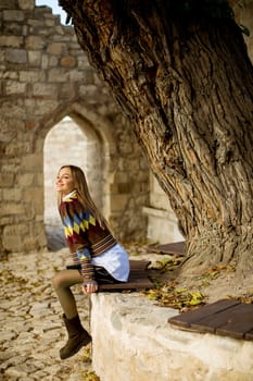 Pretty young woman sitting on the bench watching yellow leaves tree in the autumn park