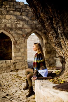 Pretty young woman sitting on the bench watching yellow leaves tree in the autumn park