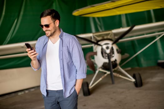 Young handsome young pilot checking airplane in the hangar and using mobile phone