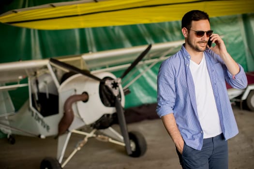 Young handsome young pilot checking airplane in the hangar and using mobile phone
