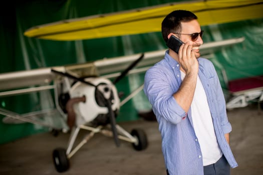 Young handsome young pilot checking airplane in the hangar and using mobile phone