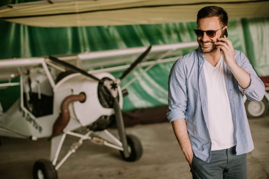 Young handsome young pilot checking airplane in the hangar and using mobile phone
