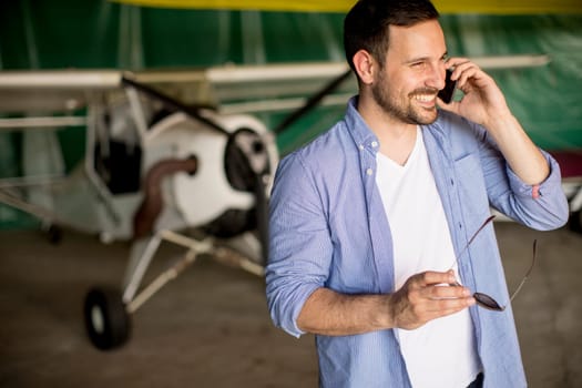Young handsome young pilot checking airplane in the hangar and using mobile phone