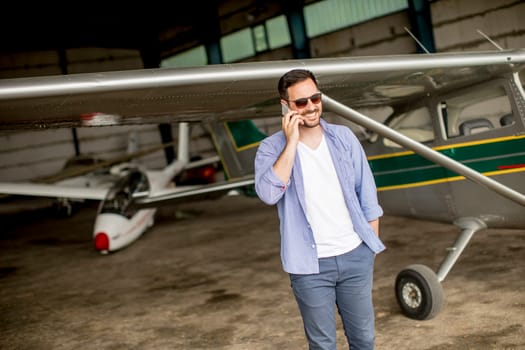 Young handsome young pilot checking airplane in the hangar and using mobile phone