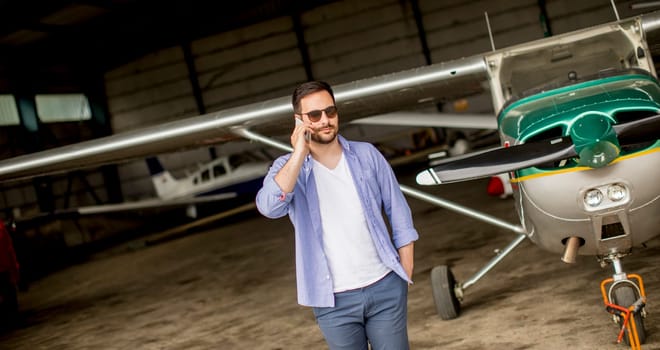 Young handsome young pilot checking airplane in the hangar and using mobile phone