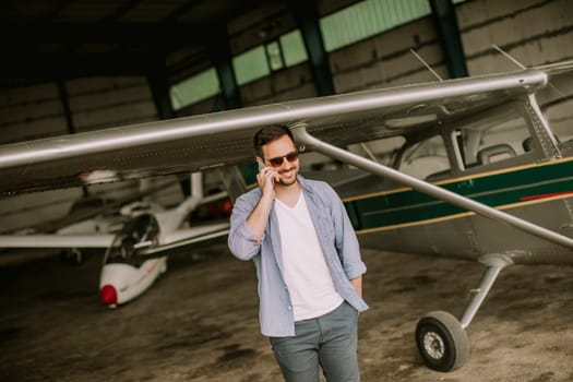 Young handsome young pilot checking airplane in the hangar and using mobile phone