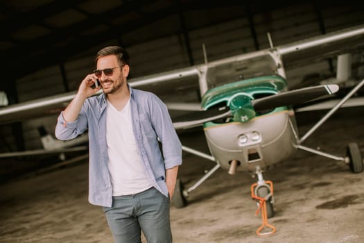 Young handsome young pilot checking airplane in the hangar and using mobile phone