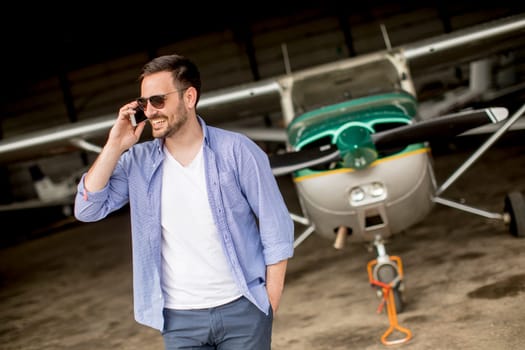 Young handsome young pilot checking airplane in the hangar and using mobile phone