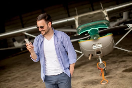 Young handsome young pilot checking airplane in the hangar and using mobile phone
