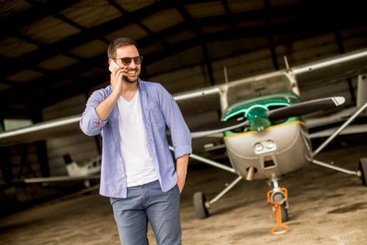 Young handsome young pilot checking airplane in the hangar and using mobile phone