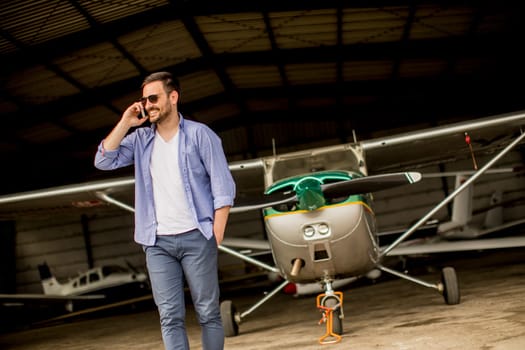Young handsome young pilot checking airplane in the hangar and using mobile phone