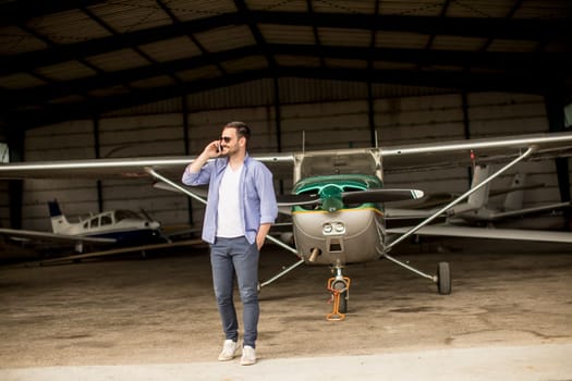 Young handsome young pilot checking airplane in the hangar and using mobile phone