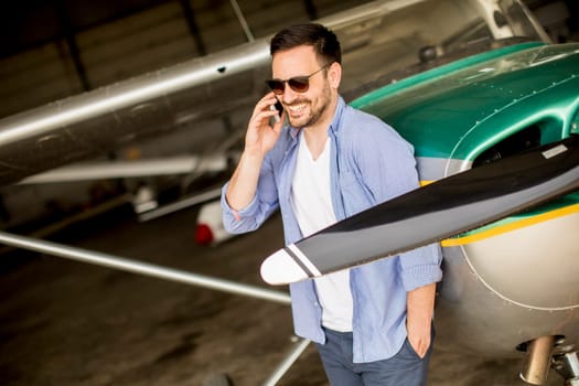 Young handsome young pilot checking airplane in the hangar and using mobile phone