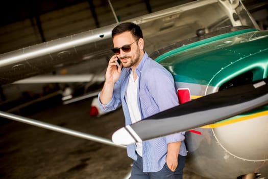 Young handsome young pilot checking airplane in the hangar and using mobile phone