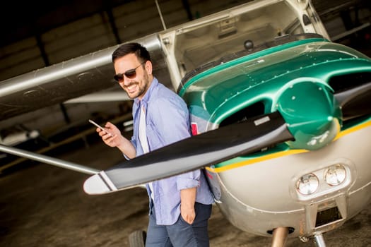 Young handsome young pilot checking airplane in the hangar and using mobile phone