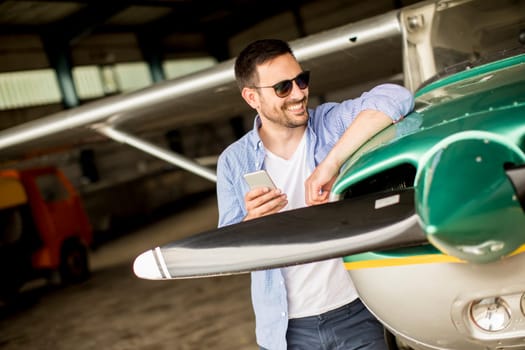 Young handsome young pilot checking airplane in the hangar and using mobile phone
