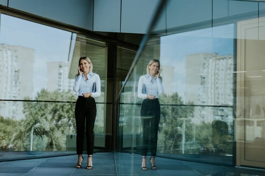 Businesswoman stands near glass windows in a contemporary office, engaged in a phone call. Urban buildings and greenery reflect in the glass.