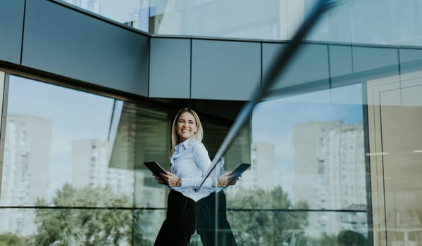 Confident businesswoman stands outside a modern glass office building, holding a tablet and smiling with a cityscape reflected in the glass