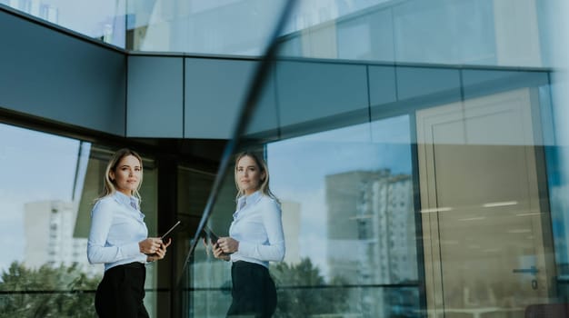 Confident businesswoman stands outside a modern glass office building, holding a tablet and smiling with a cityscape reflected in the glass