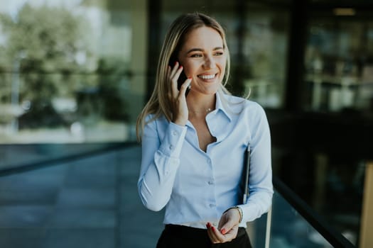 A young businesswoman smiles confidently while talking on her phone inside a sleek, modern office building, with cityscape reflections in the glass.
